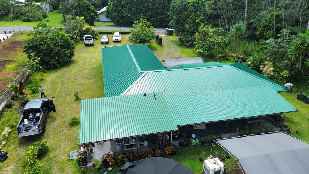 Aerial view of a green-roofed house surrounded by lush greenery and parked vehicles.