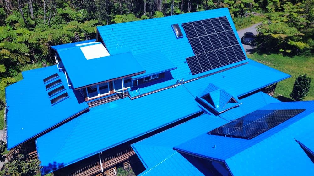 Blue metal roof with solar panels on a modern house surrounded by greenery.