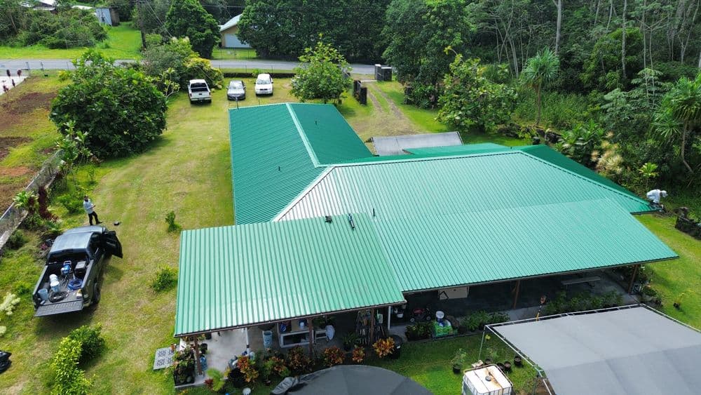 Aerial view of a green-roofed house surrounded by lush vegetation and parked vehicles.